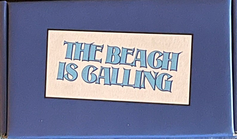 Peut inclure: Une pancarte bleue et blanche avec le texte "The Beach is Calling" sur un fond beige clair.