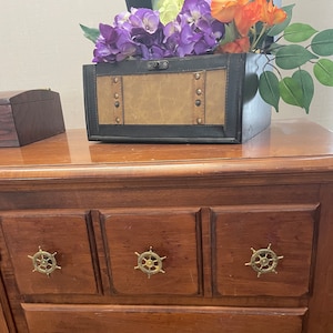 May include: A wooden dresser with three drawers, each with a gold-colored nautical wheel knob. The dresser is made of dark wood and has a light brown top. A decorative box with a leather-like finish sits on top of the dresser, with a bouquet of purple and white flowers.