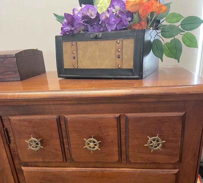 Peut inclure: Une commode en bois avec trois tiroirs, chacun avec une poign&eacute;e en forme de roue de bateau de couleur dor&eacute;e. La commode est en bois fonc&eacute; et a un dessus brun clair. Une bo&icirc;te d&eacute;corative avec une finition en cuir se trouve sur le dessus de la commode, avec un bouquet de fleurs violettes et blanches.