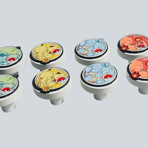May include: A set of eight round white knobs featuring colorful illustrations of various cartoon creatures. Each knob displays a unique character in a circular frame with a small red and white ball icon, set against a solid light gray background.