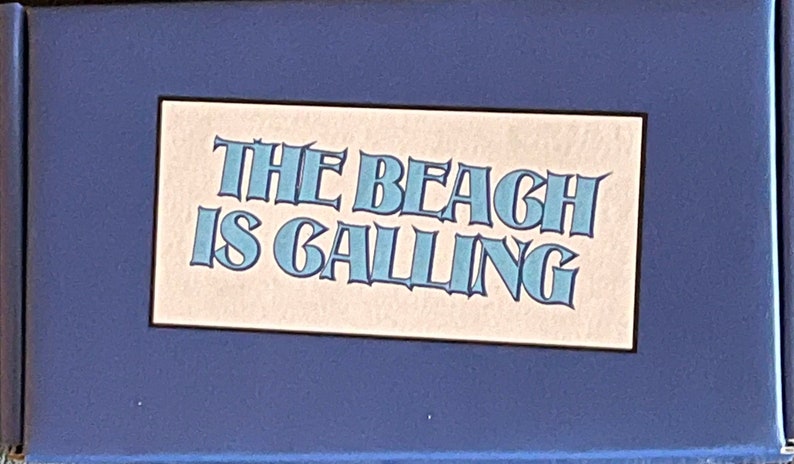 May include: A blue box with a white rectangle in the centre. The rectangle has the text "THE BEACH IS CALLING" in blue letters.