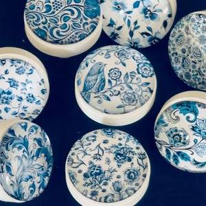 May include: A collection of seven round cabinet knobs featuring white bases and glass tops with various blue and white patterns, including floral motifs, swirling botanical designs, and a bird perched on a flowering branch.