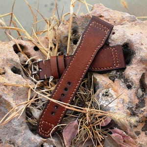 May include: A brown leather watch strap with stitching detail. The strap is lying on a rock with dry grass.