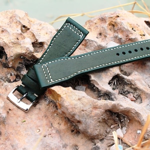 Può includere: Un cinturino da orologio in pelle verde scuro con cuciture bianche. Il cinturino ha una fibbia ed è mostrato su una superficie rocciosa.