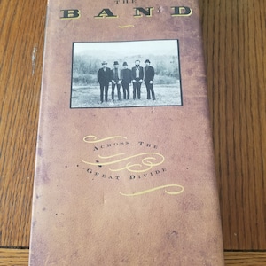 Puede incluir: El set de caja de The Band, "Across the Great Divide", presenta una foto en blanco y negro de los miembros de la banda en la portada. El set de caja es marrón con una superficie texturizada.