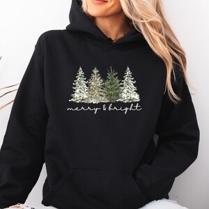 Könnte beinhalten: Schwarzer Kapuzenpullover mit einer Grafik von drei Weihnachtsbäumen in Grün-, Weiß- und Goldtönen. Der Text "merry & bright" ist in einer Schreibschrift unter den Bäumen geschrieben. Der Pullover hat eine Vordertasche.