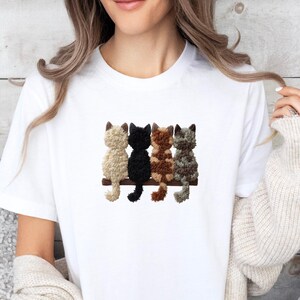 Könnte beinhalten: Weißes T-Shirt mit einem gestickten Design von vier Katzen, die auf einem braunen Holzbalken sitzen. Die Katzen sind beige, schwarz, braun und grau. Das Wort "Printed" befindet sich in einem rosa Rechteck unten rechts.