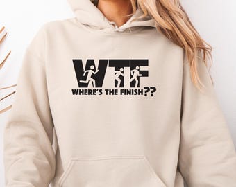 May include: A beige hoodie with a black graphic that reads "WTF Where's the Finish??" The graphic features running figures. The hoodie has a front pocket and a hood with a drawstring. The model is wearing light blue jeans.
