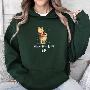 May include: A dark green hoodie with a Winnie the Pooh graphic and the text "Mama Bear To Be" and baby footprints. The graphic is in the centre of the hoodie. The hoodie has a hood and a front pocket.
