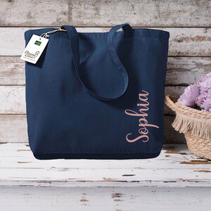 Puede incluir: Un bolso tote azul marino con el nombre "Sophia" en escritura oro rosa. El bolso tiene asas largas y una etiqueta "Organic Cotton". A la derecha hay una cesta tejida con flores moradas.