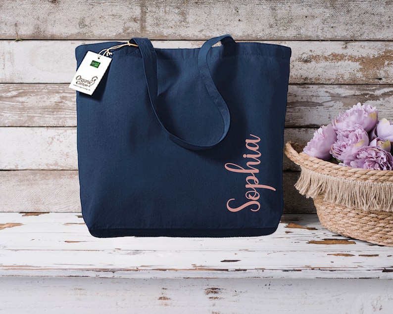 Puede incluir: Un bolso tote azul marino con el nombre "Sophia" en escritura oro rosa. El bolso tiene asas largas y una etiqueta "Organic Cotton". A la derecha hay una cesta tejida con flores moradas.