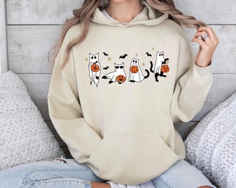 May include: A beige hoodie with a Halloween design. The graphic shows five cartoon cats in ghost costumes, each carrying a pumpkin. The design also includes black bats and gold stars. The hoodie has a front pocket and a hood.