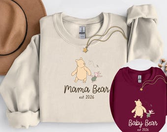 May include: A cream-coloured crewneck sweatshirt featuring a graphic of Winnie the Pooh and Piglet with the text "mama Bear est 2026" and a maroon sweatshirt with the same graphic and the text "Baby Bear est 2026", both paired with a gold star necklace.