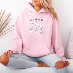 May include: A pale pink hoodie with a front pocket and a drawstring hood. The hoodie displays a graphic of cartoon geese with the text "it is okay to" above them. The geese have various accessories and speech bubbles.