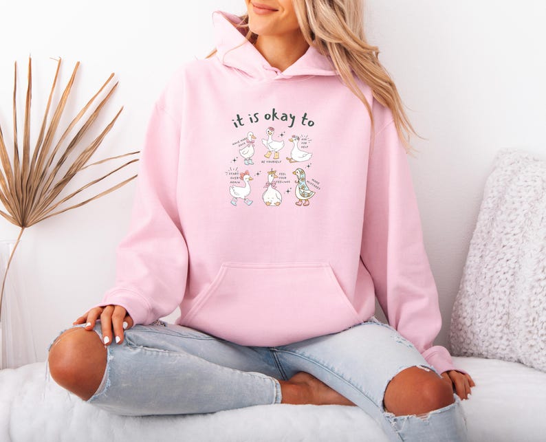 May include: A pale pink hoodie with a front pocket and a drawstring hood. The hoodie displays a graphic of cartoon geese with the text "it is okay to" above them. The geese have various accessories and speech bubbles.