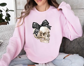 May include: A light pink long-sleeve sweatshirt featuring a graphic print of a human skull wearing a large black and grey leopard print bow on its head. The sweatshirt has a crew neckline and long sleeves with ribbed cuffs and a ribbed hem.