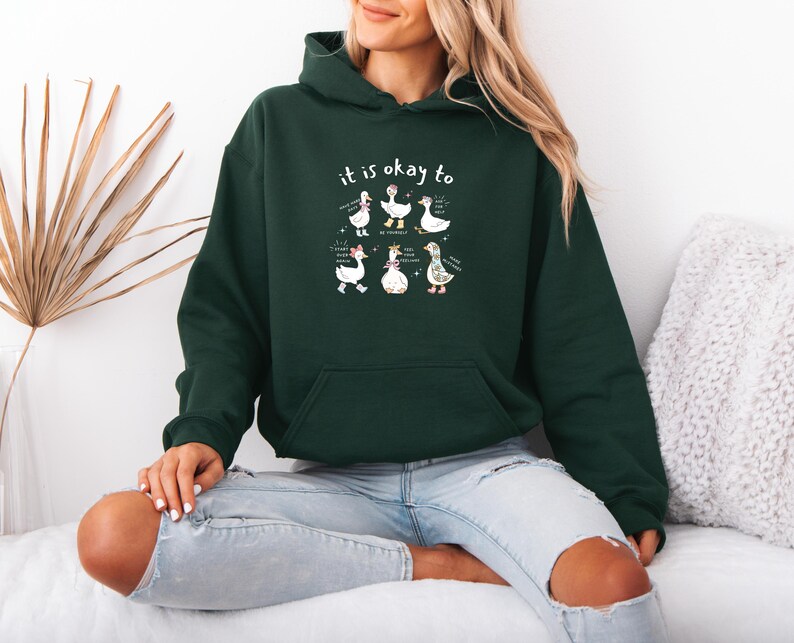 May include: Dark green hoodie with a front pocket and a hood. The hoodie features the text "it is okay to" above a graphic of six cartoon geese. The geese are in various poses with text bubbles.