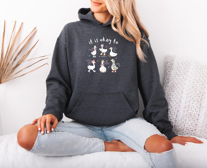 May include: Dark grey hoodie with a front pocket and a drawstring hood. The hoodie features a graphic of cartoon geese with the text "it is okay to" above them. The geese are in various poses with text below them.