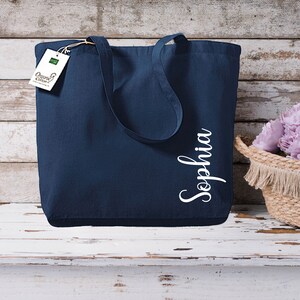 Puede incluir: Bolso tote azul marino con el nombre "Sophia" en escritura blanca. El bolso tiene asas largas y una etiqueta que dice "Organic Cotton". A la derecha hay una cesta tejida con flores moradas.