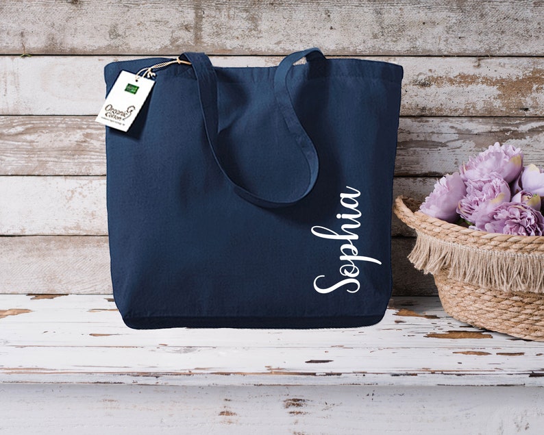 Puede incluir: Bolso tote azul marino con el nombre "Sophia" en escritura blanca. El bolso tiene asas largas y una etiqueta que dice "Organic Cotton". A la derecha hay una cesta tejida con flores moradas.