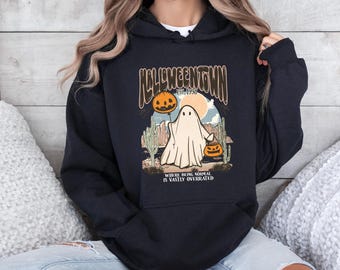 May include: Black Halloween-themed hoodie with a graphic featuring a ghost holding a jack-o'-lantern, a pumpkin balloon, and the text "Halloweentown Est. 1998". The design includes cacti and the phrase "Where being normal is vastly overrated."