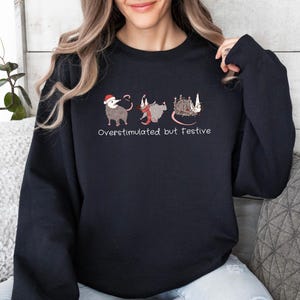 May include: A black long-sleeve sweatshirt featuring a graphic of three opossums in holiday attire and the text "Overstimulated but Festive" printed in white. The first opossum wears a Santa hat, the second a red scarf, and the third is tangled in lights.
