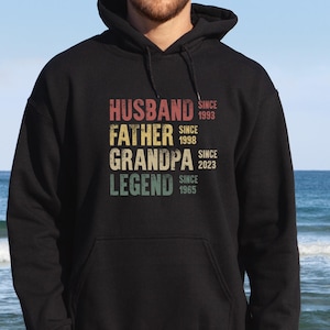 Könnte beinhalten: Schwarzer Kapuzenpullover mit den Worten "HUSBAND, FATHER, GRANDPA, LEGEND" in einem Vintage-Schriftstil. Der Pullover enthält auch die Jahreszahlen "SINCE 1993, SINCE 1998, SINCE 2023, SINCE 1965".