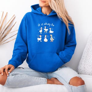 May include: A royal blue hoodie with a front pocket and a drawstring hood. The hoodie features the text "it is okay to" above a graphic of cartoon geese. The geese are in various poses with text bubbles.