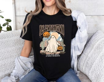 May include: Black t-shirt with a Halloween-themed graphic. The design features a ghost, pumpkins, and the text "Halloweentown Est. 1998". The phrase "Where being normal is vastly overrated" is also included.