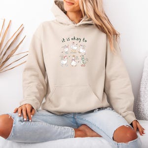 May include: A beige hooded sweatshirt with a front pocket. The design includes the words "it is okay to" and a cartoon illustration of geese with accessories and speech bubbles. The geese are in different poses.
