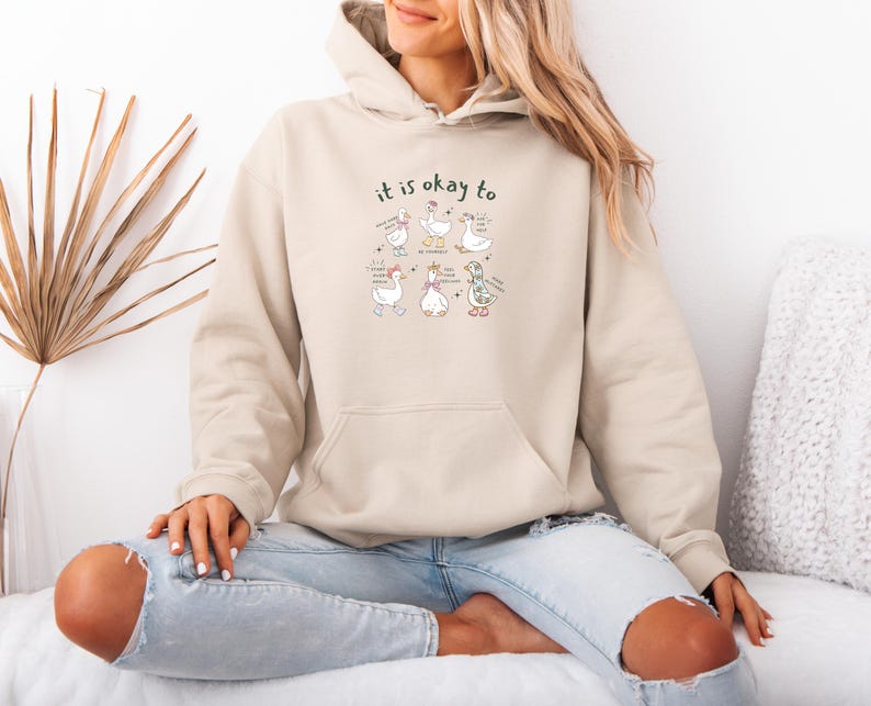 May include: A beige hooded sweatshirt with a front pocket. The design includes the words "it is okay to" and a cartoon illustration of geese with accessories and speech bubbles. The geese are in different poses.