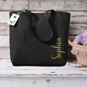 Puede incluir: Bolso tote negro con el nombre "Sophia" en letras doradas. El bolso tiene asas largas y una etiqueta que dice "Organic Cotton". Una cesta de mimbre con flores moradas est&aacute; cerca.