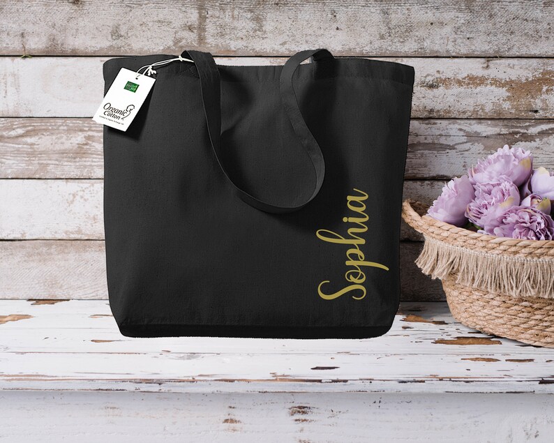 Puede incluir: Bolso tote negro con el nombre "Sophia" en letras doradas. El bolso tiene asas largas y una etiqueta que dice "Organic Cotton". Una cesta de mimbre con flores moradas est&aacute; cerca.