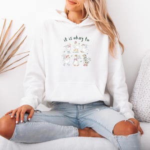 May include: White hooded sweatshirt featuring a front pocket and the words "it is okay to" above a graphic of cartoon geese. The geese are in different poses with speech bubbles. The sweatshirt is being worn.