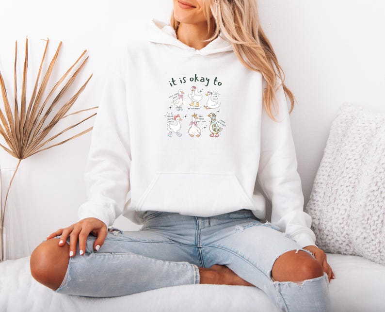 May include: White hooded sweatshirt featuring a front pocket and the words "it is okay to" above a graphic of cartoon geese. The geese are in different poses with speech bubbles. The sweatshirt is being worn.