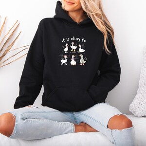 May include: Black hoodie featuring the phrase "it is okay to" and a graphic of six cartoon geese. The geese are depicted in different poses and outfits. The hoodie has a front pocket and a drawstring hood.