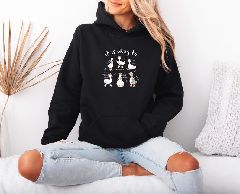 May include: Black hoodie featuring the phrase "it is okay to" and a graphic of six cartoon geese. The geese are depicted in different poses and outfits. The hoodie has a front pocket and a drawstring hood.
