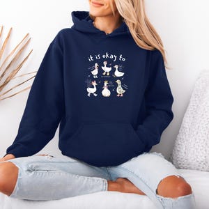 May include: A navy blue hooded sweatshirt featuring the words "it is okay to" above a cartoon illustration of six geese. Each goose has a unique design. The sweatshirt has a front pocket and a drawstring hood.