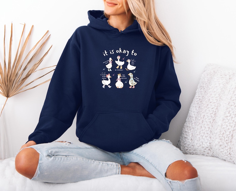 May include: A navy blue hooded sweatshirt featuring the words "it is okay to" above a cartoon illustration of six geese. Each goose has a unique design. The sweatshirt has a front pocket and a drawstring hood.