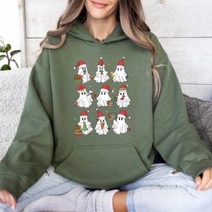 May include: An olive green hoodie with a graphic of cartoon ghosts wearing Santa hats and holding books. The ghosts are white with black eyes and are arranged in a grid pattern. The hoodie has a kangaroo pocket.