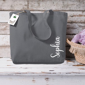 Puede incluir: Un bolso tote gris con el nombre "Sophia" en escritura blanca. El bolso tiene asas largas y una etiqueta que dice "Organic Cotton". Una cesta tejida con flores moradas est&aacute; a la derecha.