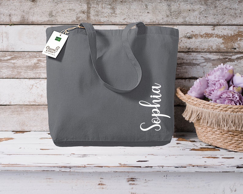 Puede incluir: Un bolso tote gris con el nombre "Sophia" en escritura blanca. El bolso tiene asas largas y una etiqueta que dice "Organic Cotton". Una cesta tejida con flores moradas est&aacute; a la derecha.