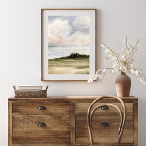 May include: A watercolour painting of a landscape with a cloudy sky and a green hill in the distance. The painting is framed in a light wood frame and is hanging on a white wall.