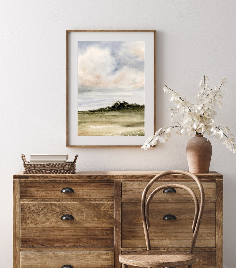 May include: A watercolour painting of a landscape with a cloudy sky and a green hill in the distance. The painting is framed in a light wood frame and is hanging on a white wall.