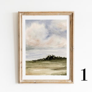 May include: A framed watercolour painting of a landscape with a cloudy sky and a green hill in the foreground. The sky is a soft blue with pink and grey clouds. The hill is a dark green with a few trees on top. The painting is in a light wood frame.