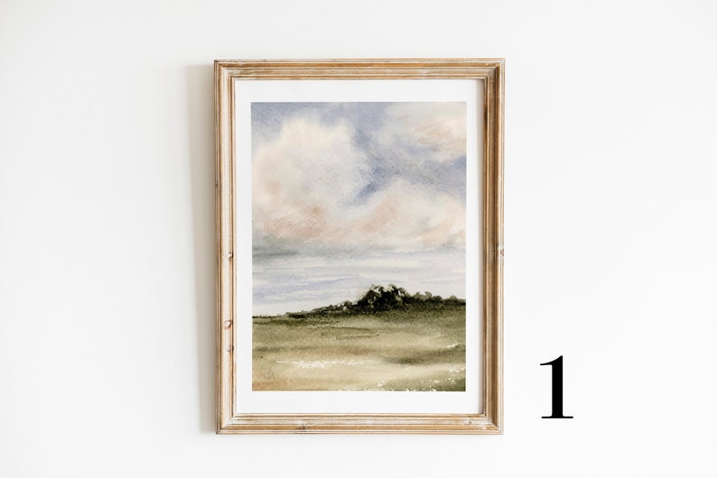 May include: A framed watercolour painting of a landscape with a cloudy sky and a green hill in the foreground. The sky is a soft blue with pink and grey clouds. The hill is a dark green with a few trees on top. The painting is in a light wood frame.