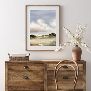 May include: A watercolour painting of a landscape with a cloudy sky, green hills, and a body of water. The painting is framed in a light wood frame and is hanging on a white wall.
