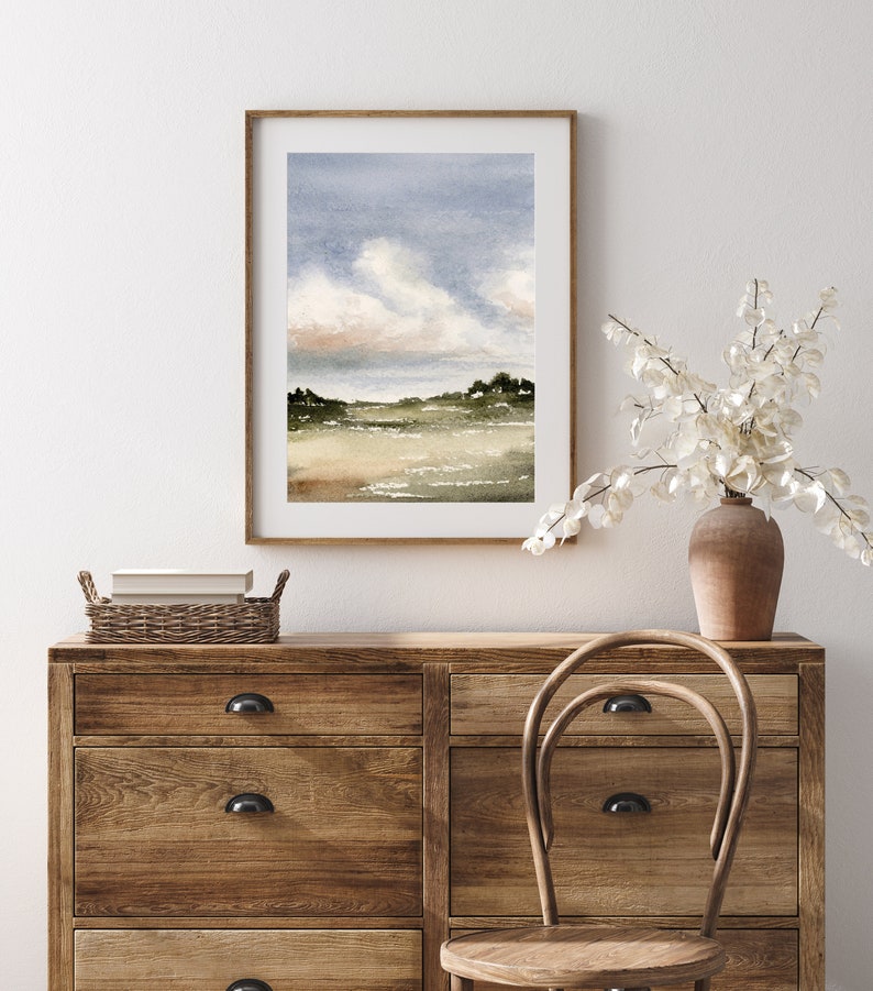 May include: A watercolour painting of a landscape with a cloudy sky, green hills, and a body of water. The painting is framed in a light wood frame and is hanging on a white wall.
