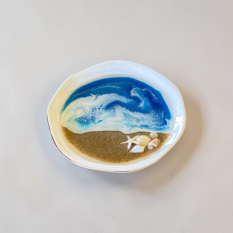 May include: A white, irregularly shaped tray featuring a resin art design that mimics a beach scene. The surface displays a sandy brown base with a blue and white wave pattern, accented by a small white starfish and several miniature seashells.