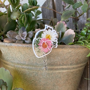 May include: Clear acrylic badge reel shaped like a heart, containing dried flowers in pink, white, and yellow. The reel has a silver retractable cord and a clear plastic clip. The flowers are measured in centimeters.